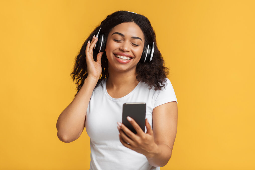 Modern technologies. African american lady listening music in wireless headphones, searching song on smartphone over yellow background. Black woman enjoying audio playlist
